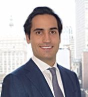 Headshot of a young man with dark hair in a suit, city buildings behind